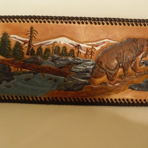 May include: A brown leather wallet with a carved image of a bear walking through a mountain landscape. The bear is brown and the mountains are white with a blue river in the foreground.