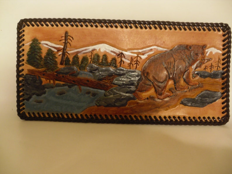May include: A brown leather wallet with a carved image of a bear walking through a mountain landscape. The bear is brown and the mountains are white with a blue river in the foreground.
