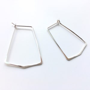 May include: A pair of silver-colored geometric earrings. Each earring has a unique, angular shape with a flat bottom and a slightly textured surface. The earrings are displayed against a white background.