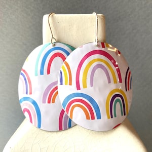 May include: A pair of round earrings with a white background and a repeating rainbow pattern in various colors. The earrings have gold-colored hooks.