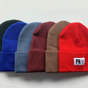 May include: Six knit beanies in different colors, including black, blue, gray, burgundy, brown, and red. The red beanie has a white patch with the letters "PN" and a peace sign.
