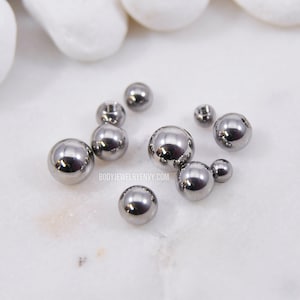 May include: A collection of small, polished, gunmetal-colored spherical beads of varying sizes. The text BODYJEWELRYENVY.COM is printed in small white letters among the beads. These metallic spheres have a reflective surface and a smooth finish.