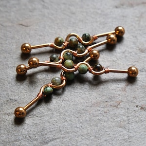 May include: Three industrial barbell body jewelry pieces featuring a copper-toned metal bar with a central wavy design. Each bar is adorned with small, round, mottled green and brown stone beads wrapped in thin copper wire, ending in smooth metal balls.