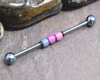 May include: A straight industrial barbell body jewelry piece featuring a dark metallic grey shaft with two matching spherical end caps. The center of the barbell is adorned with three small beads in light blue, bright pink, and light pink colors.