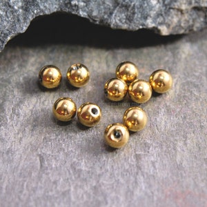 May include: A collection of small, spherical, gold-colored metal beads with a polished, reflective surface. Each bead features a single, centered circular hole. The beads are arranged in a loose cluster, showcasing their smooth, metallic finish.