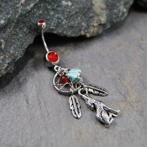 May include: A silver-toned belly button ring featuring a curved barbell with a red crystal top and bottom ball. A dangling charm assembly includes a circular dreamcatcher frame, a turquoise stone, a red bead, two silver feathers, and a wolf.