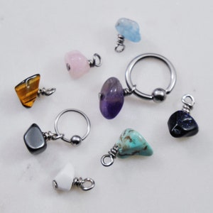 May include: A collection of jewelry components including two circular metal rings and several tumbled gemstone charms. The stones include tiger's eye, rose quartz, amethyst, turquoise, white stone, and dark blue glitter stone, each attached to a metal loop.