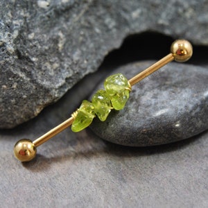May include: A gold-toned industrial barbell body jewelry piece featuring three small, raw, translucent lime green peridot gemstone chips wrapped securely around the center of the straight metal bar with thin gold-colored wire.