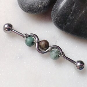 May include: A silver barbell with a green and brown stone bead on each end. The barbell is on a white surface.