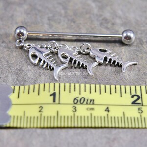 May include: A silver-toned industrial barbell body jewelry piece featuring three dangling fish skeleton charms. A yellow measuring tape is positioned below the jewelry, showing the barbell length is approximately 1.5 inches long.