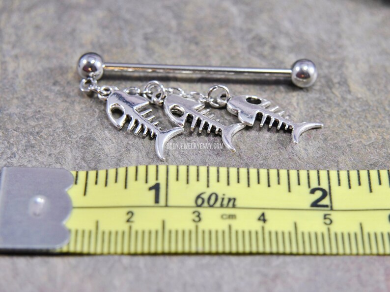 May include: A silver-toned industrial barbell body jewelry piece featuring three dangling fish skeleton charms. A yellow measuring tape is positioned below the jewelry, showing the barbell length is approximately 1.5 inches long.