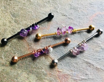 May include: Four industrial barbell piercings featuring straight metal bars in black, gold, rose gold, and silver tones. Each barbell is adorned with a cluster of raw, purple amethyst stones secured to the center of the bar with thin wire wrapping.