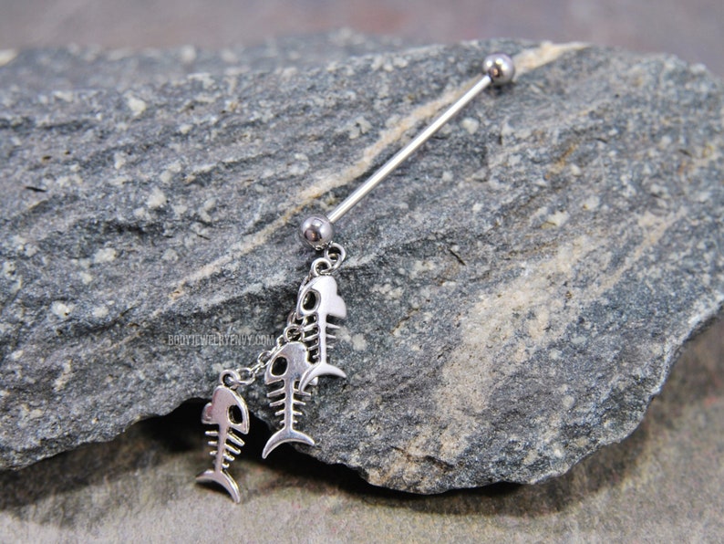 May include: A silver industrial barbell earring featuring three fish skeleton charms. The barbell has a ball on each end, and the charms dangle from a chain. The earring is displayed on a gray stone surface.
