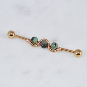 May include: A gold-toned industrial barbell body jewelry piece featuring a central wire-wrapped section with three small, round, mottled green and black stone beads. The barbell has smooth, rounded end caps and the text BODYJEWELRYENVY.COM is visible.