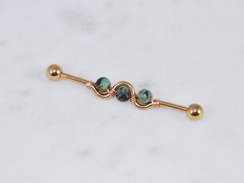 May include: A gold-toned industrial barbell body jewelry piece featuring a central wire-wrapped section with three small, round, mottled green and black stone beads. The barbell has smooth, rounded end caps and the text BODYJEWELRYENVY.COM is visible.