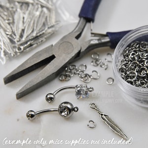 Puede incluir: Dos piezas de joyer&iacute;a corporal tipo barra curva con piedras transparentes, un dije de pluma plateado y varios anillos de salto met&aacute;licos peque&ntilde;os. Se ven unos alicates de punta con mangos azul oscuro y un recipiente con anillos.