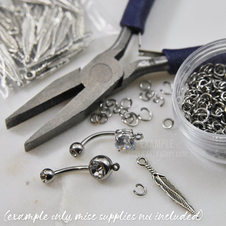 Puede incluir: Dos piezas de joyer&iacute;a corporal tipo barra curva con piedras transparentes, un dije de pluma plateado y varios anillos de salto met&aacute;licos peque&ntilde;os. Se ven unos alicates de punta con mangos azul oscuro y un recipiente con anillos.