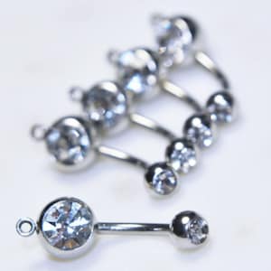 Puede incluir: Una colecci&oacute;n de joyas corporales de barra curva en tono plateado con cristales circulares transparentes y facetados. Cada pieza consta de un engaste de cristal m&aacute;s grande con una peque&ntilde;a anilla y una bola con cristal en el extremo.