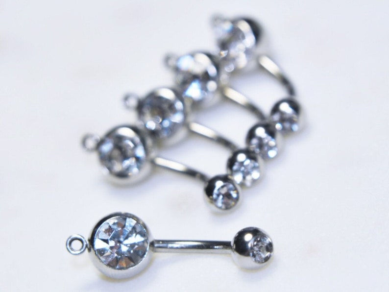 Puede incluir: Una colecci&oacute;n de joyas corporales de barra curva en tono plateado con cristales circulares transparentes y facetados. Cada pieza consta de un engaste de cristal m&aacute;s grande con una peque&ntilde;a anilla y una bola con cristal en el extremo.