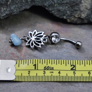 May include: A silver-toned belly button ring featuring a lotus flower charm, a clear round gemstone, and a light blue tumbled stone bead. The jewelry is positioned above a yellow measuring tape showing a length of approximately 1.5 inches.