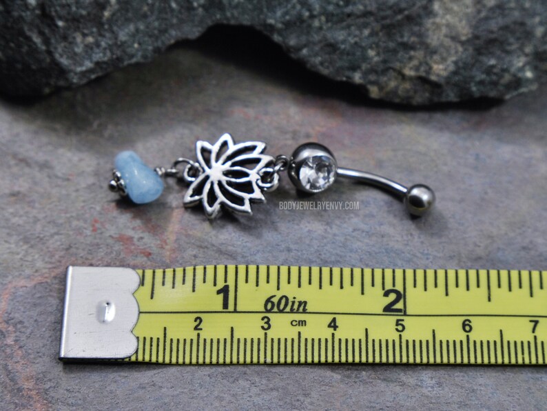 May include: A silver-toned belly button ring featuring a lotus flower charm, a clear round gemstone, and a light blue tumbled stone bead. The jewelry is positioned above a yellow measuring tape showing a length of approximately 1.5 inches.
