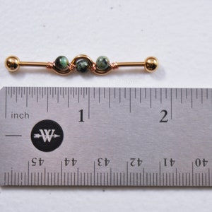 May include: A gold-toned industrial barbell body jewelry piece featuring three small green and black speckled stone beads wrapped in copper-colored wire. The barbell measures approximately 1.5 inches in length as shown by the metal ruler.