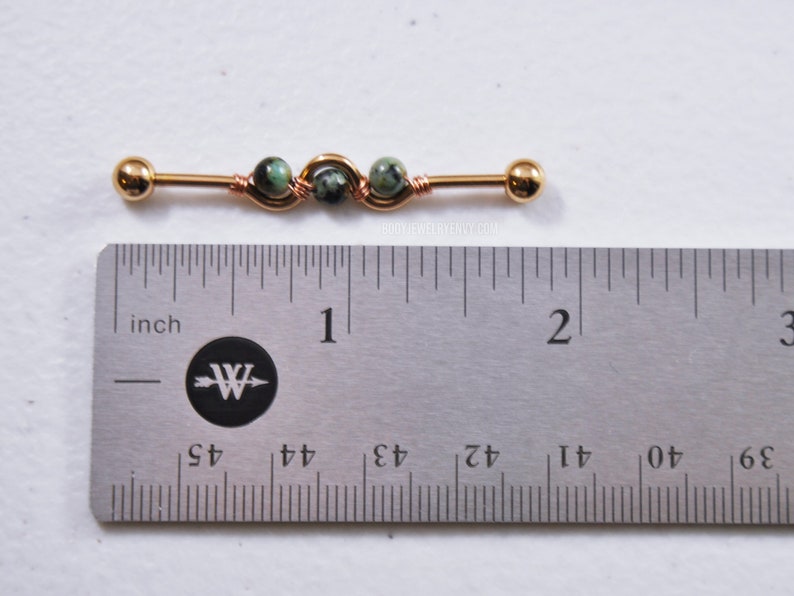 May include: A gold-toned industrial barbell body jewelry piece featuring three small green and black speckled stone beads wrapped in copper-colored wire. The barbell measures approximately 1.5 inches in length as shown by the metal ruler.