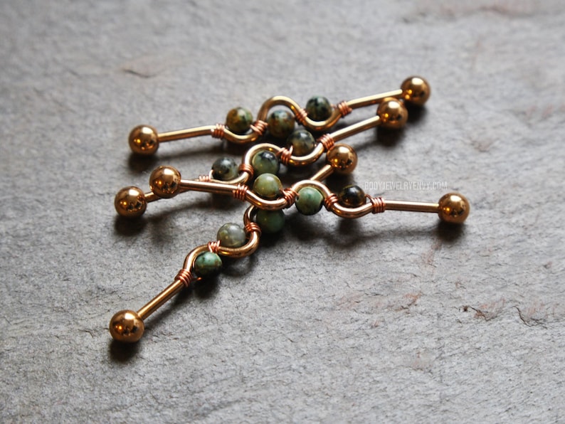 May include: Three industrial barbell body jewelry pieces featuring a copper-toned metal bar with a central wavy design. Each bar is adorned with small, round, mottled green and brown stone beads wrapped in thin copper wire, ending in smooth metal balls.