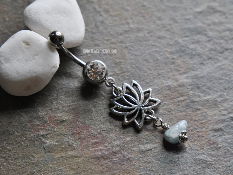 May include: A silver-toned curved barbell belly ring featuring a clear round gemstone, a dangling silver lotus flower charm, and a small light blue stone bead at the end. The text BODYJEWELRYENVY.COM is printed in small white letters near the top.