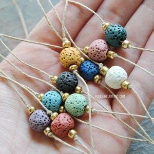 Puede incluir: Un conjunto de 10 pulseras de piedra de lava con cuentas de diferentes colores. Cada pulsera está hecha con una cuenta de piedra de lava natural, una cuenta dorada y un cordón fino de cuerda natural. Las pulseras están dispuestas en una mano, mostrando los diferentes colores y estilos.