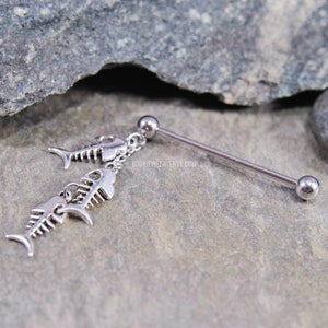 May include: A silver-toned industrial barbell body jewelry piece featuring a dangling chain with two fish skeleton charms. The barbell has a smooth metal shaft with a ball at each end. The text BODYJEWELRYENVY.COM is visible on the metal surface.