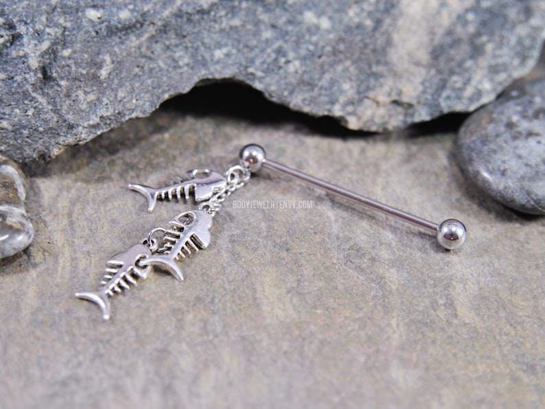 May include: A silver-toned industrial barbell body jewelry piece featuring a dangling chain with two fish skeleton charms. The barbell has a smooth metal shaft with a ball at each end. The text BODYJEWELRYENVY.COM is visible on the metal surface.