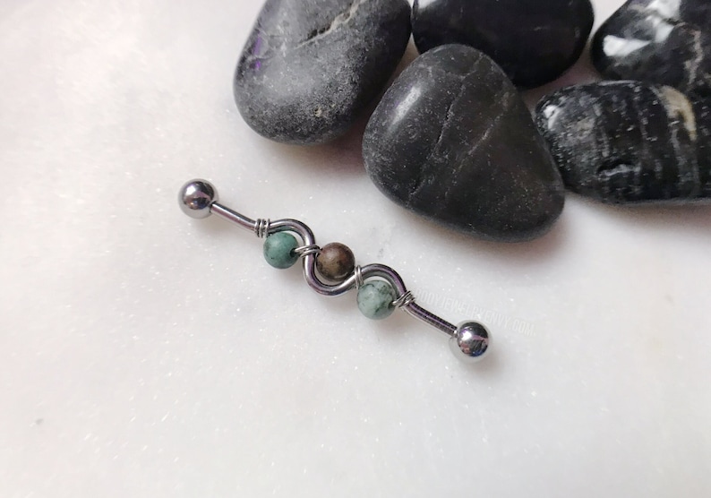May include: A silver barbell with a green and brown stone bead on each end. The barbell is on a white surface.