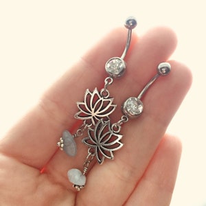 May include: Two silver-toned curved barbell navel rings featuring a clear round crystal at the top, a dangling open-work lotus flower charm in the center, and a small, irregular, pale blue-grey stone bead at the bottom of each piece.