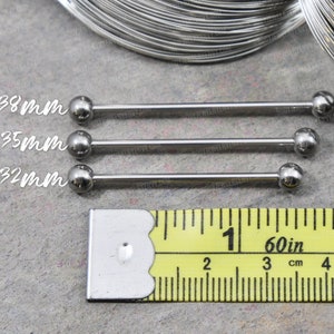 May include: Three silver-colored metal industrial barbell piercings with spherical ends. The text 14g 38mm, 14g 35mm, and 14g 32mm is printed next to each bar. A yellow measuring tape is positioned below, showing measurements in inches and centimeters.