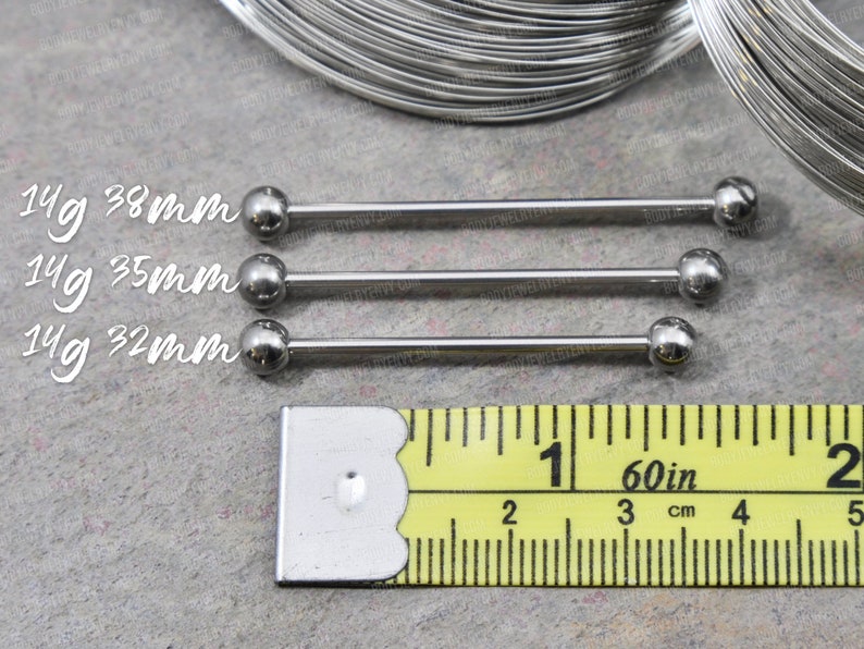 May include: Three silver-colored metal industrial barbell piercings with spherical ends. The text 14g 38mm, 14g 35mm, and 14g 32mm is printed next to each bar. A yellow measuring tape is positioned below, showing measurements in inches and centimeters.