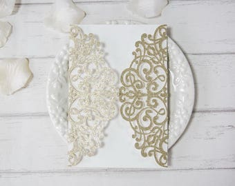 May include: Two laser cut wedding invitation envelopes with a delicate, intricate design. The envelope on the left is silver glitter and the envelope on the right is gold glitter.