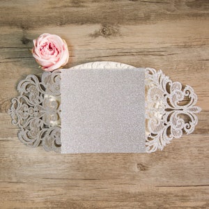 May include: A silver glitter card with a delicate white lace design. The card is open and ready to be written on.