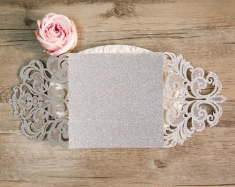 May include: A silver glitter card with a delicate white lace design. The card is open and ready to be written on.