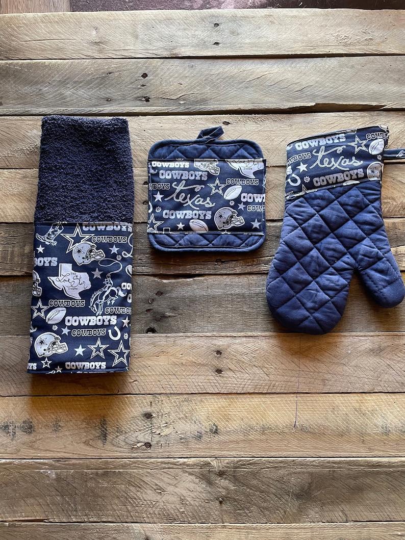 May include: A set of three kitchen towels featuring a navy blue background with white Dallas Cowboys logos, stars, and footballs. The set includes a hand towel, a pot holder, and an oven mitt.