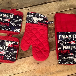Pode incluir: Um conjunto de acessórios de cozinha vermelhos e azuis com o logotipo do New England Patriots. O conjunto inclui dois pegadores, uma luva de forno e um pano de cozinha.
