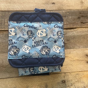 Pode incluir: Um pegador de panela azul e branco com um suporte acolchoado azul marinho. A frente apresenta um padr&atilde;o repetido do logotipo dos Tar Heels da Universidade da Carolina do Norte, o mascote e as palavras "Tar Heels" e "North Carolina".