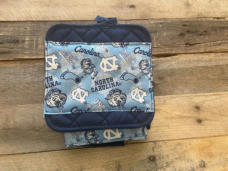 Pode incluir: Um pegador de panela azul e branco com um suporte acolchoado azul marinho. A frente apresenta um padr&atilde;o repetido do logotipo dos Tar Heels da Universidade da Carolina do Norte, o mascote e as palavras "Tar Heels" e "North Carolina".