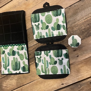 May include: A set of kitchen accessories featuring a black kitchen towel with a green cactus print border, two black pot holders with a green cactus print, and a small green cactus-shaped trivet.