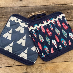 May include: Two navy blue quilted oven mitts. One features white teepee designs, the other has a pattern of colorful feathers. Both have a white scalloped trim and a navy blue hanging loop.