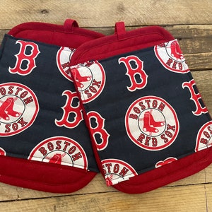May include: Two Boston Red Sox oven mitts. The mitts are navy blue with a repeating pattern of the team's logo and the letter "B" in red and white. The cuffs and hanging loops are solid red.