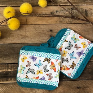 May include: Two teal quilted pot holders with a white background and a colorful butterfly pattern. The pot holders have a teal border with a geometric pattern.