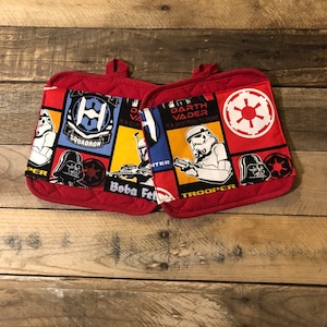 May include: Two red quilted pot holders with a Star Wars theme. The pot holders feature images of Darth Vader, Boba Fett, Stormtroopers, and the Imperial insignia.