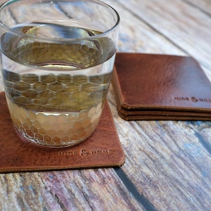 May include: A stack of square, brown leather coasters with the embossed text "HIDE & HOME". A glass of water sits on one coaster. The coasters are made of a textured, distressed leather. The image is taken on a wooden surface.