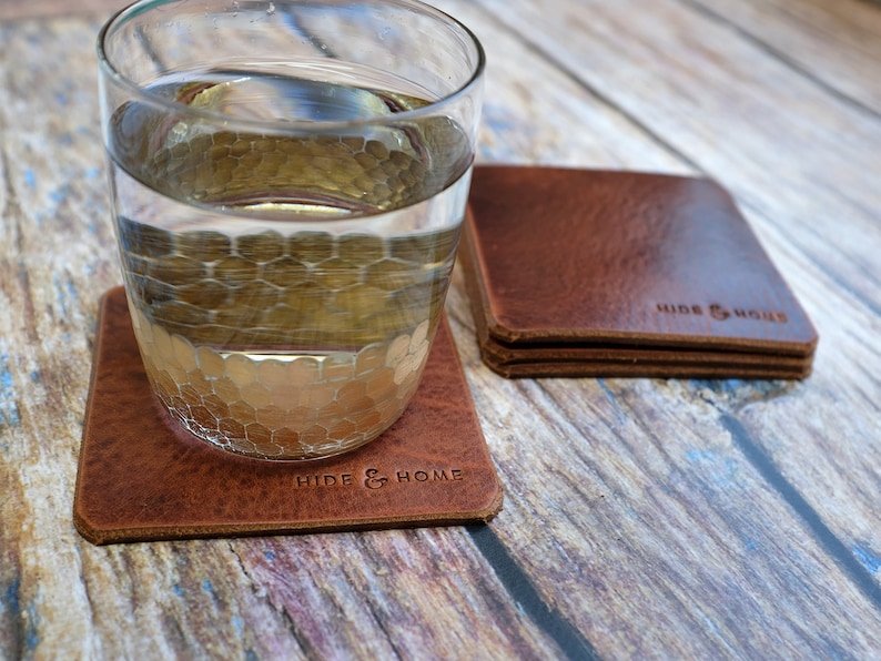 May include: A stack of square, brown leather coasters with the embossed text "HIDE & HOME". A glass of water sits on one coaster. The coasters are made of a textured, distressed leather. The image is taken on a wooden surface.